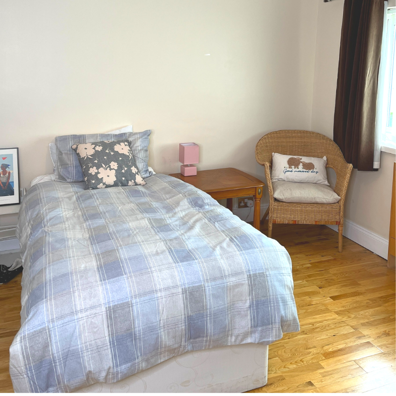 A bedroom with a comfy looking bedspread, a side table and a chair with a cushion. The side window is just visible.